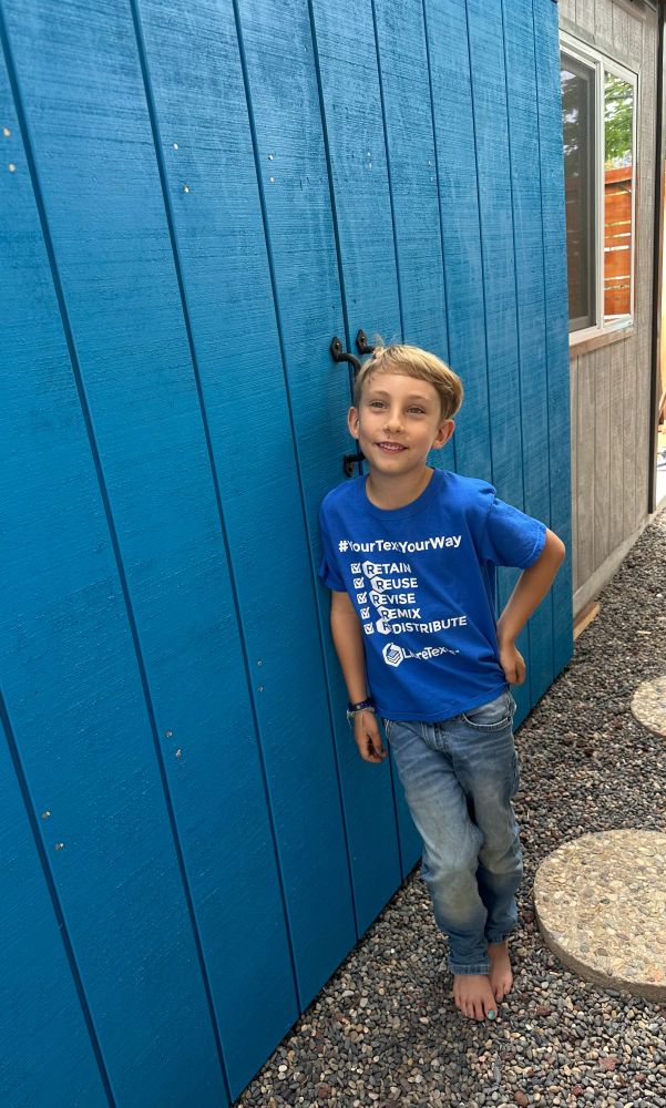 Young boy with blonde hair wearing a LibreTexts shirt and blue jeans smiling as he stands in front of a bright blue shed door