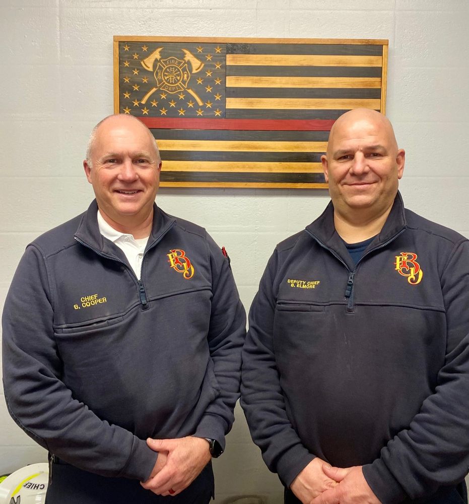 Two men in blue "BFD" sweaters stand in front of a wooden flag featuring the fire department's crest and a red line