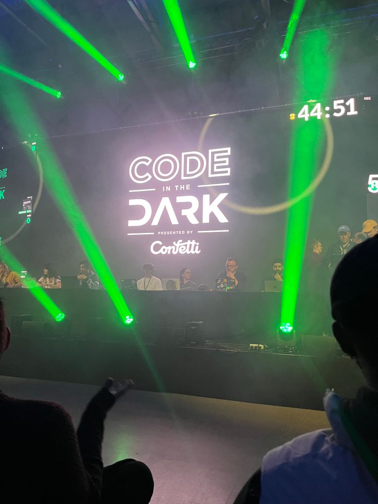 The main stage at Nordic.JS. A row of people sit at a long table at computers (I'm not among them yet) and the screen says "CODE IN THE DARK sponsored by confetti"