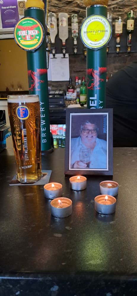 A photographer of my recently deceased dad on the bar of the village pub. The photo is of him smiling and drinking a pint. In front of the photo are five tea light candles, and there is a pint of Fosters for him on the left hand side of the image. 