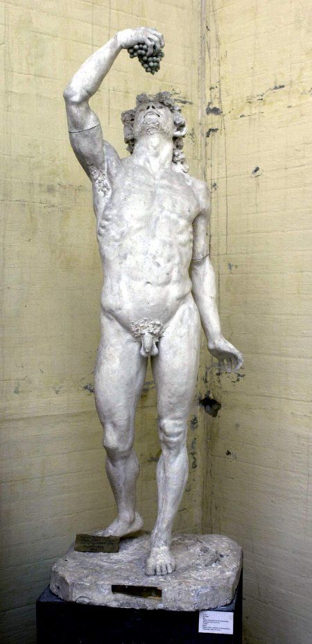 Escultura en escayola blanca de un hombre desnudo, un fauno, con el brazo levantado sobre su cabeza sosteniendo un racimo de uva. La cabeza está erguida hacia arriba mirando el racimo