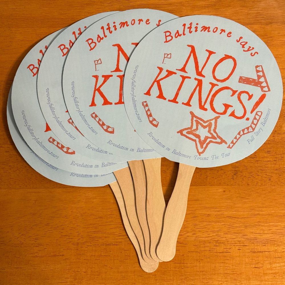Photo of five circular hand fans, pale blue and orange, that have an illustrated design and the words "Baltimore says NO KINGS!" on them.