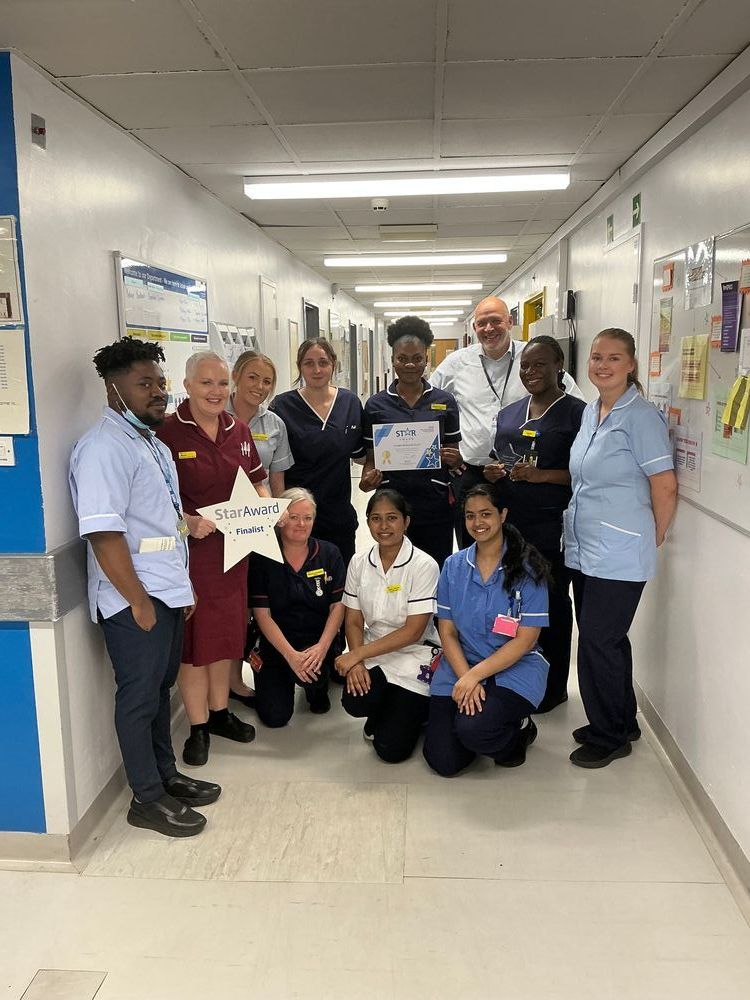 Group shot of team members from the Acute Medical Unit. They are standing together on a hospital corridor with Trust Chief Executive, Simon. One member of the team is holding a Star Award certificate, another their trophy, and another a 'Star Award finalist' star-shaped sign.