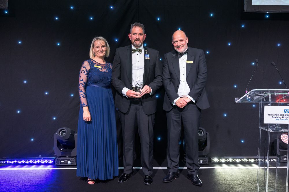 Rich Atkinson, last year's Patient Choice Award winner, onstage with Dawn Parkes, Chief Nurse, and Trust Chief Executive, Simon Morritt. All three colleagues are wearing formal clothing.