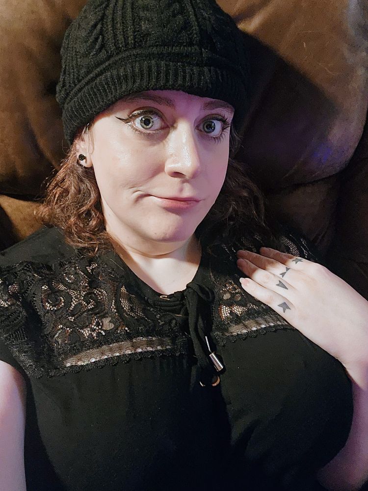 A selfie of me in a brown leather chair. I'm wearing a lacy black top and a black hat