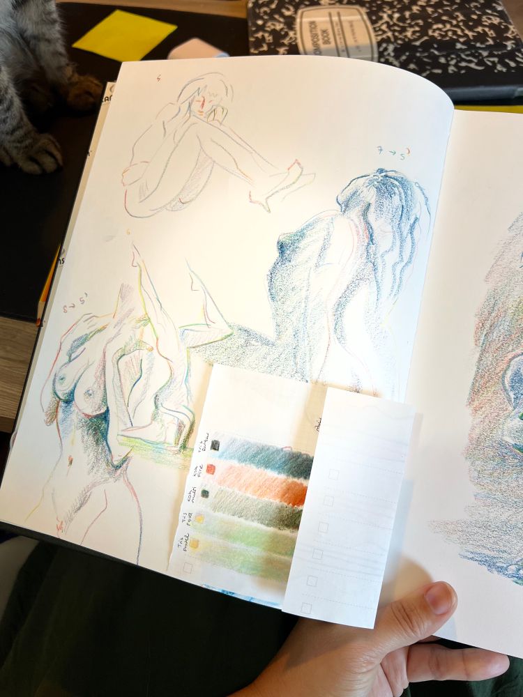 Foto de esboços de silhuetas femininas nuas feito em lápis de cor em um caderno, com um papel dobrado sobre eles em que se vêm alguns gradientes coloridos. Como na outra foto, dá pra ver no fundo alguns objetos coloridos e as patas dianteiras de um gato tigrado (a Matilda)