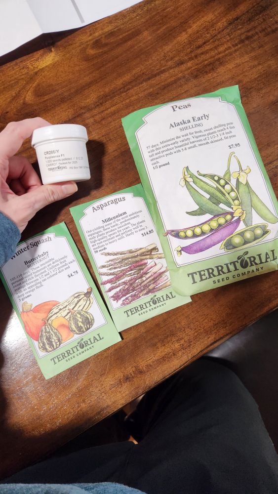 A picture of some seed packets sitting on a table. Butter baby squash, millennium asparagus, Alaska early peas, and purplesnax carrots. From territorial seed company. 