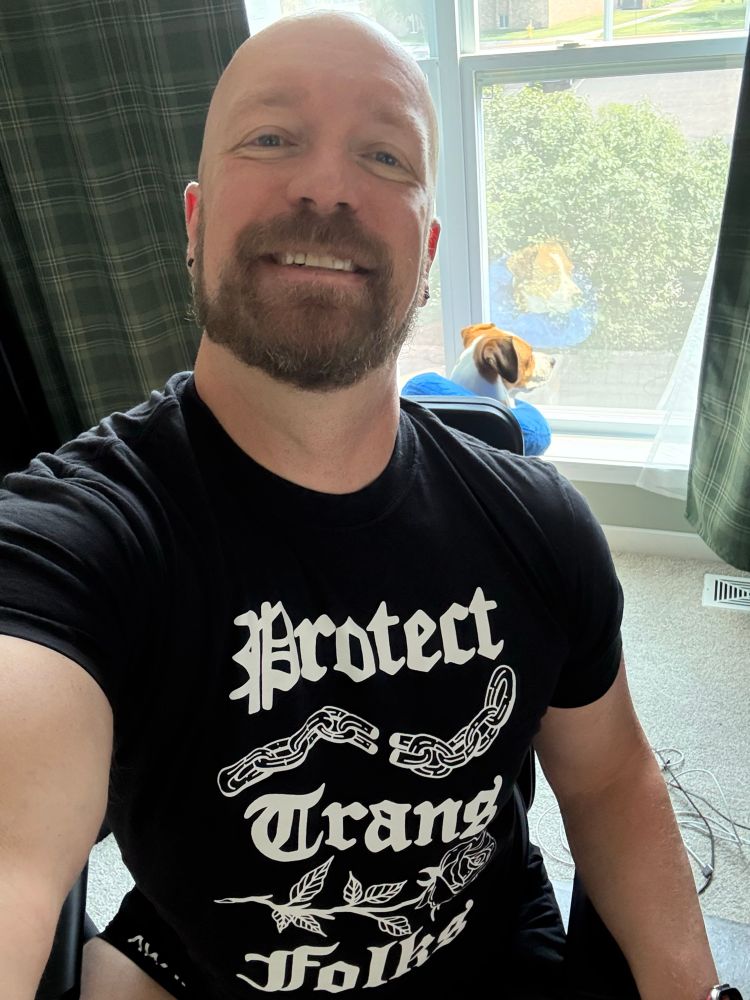 Pic of me. Bald and bearded. In a black tee that says Protect Trans Folks. Luma in the background in a donut.
