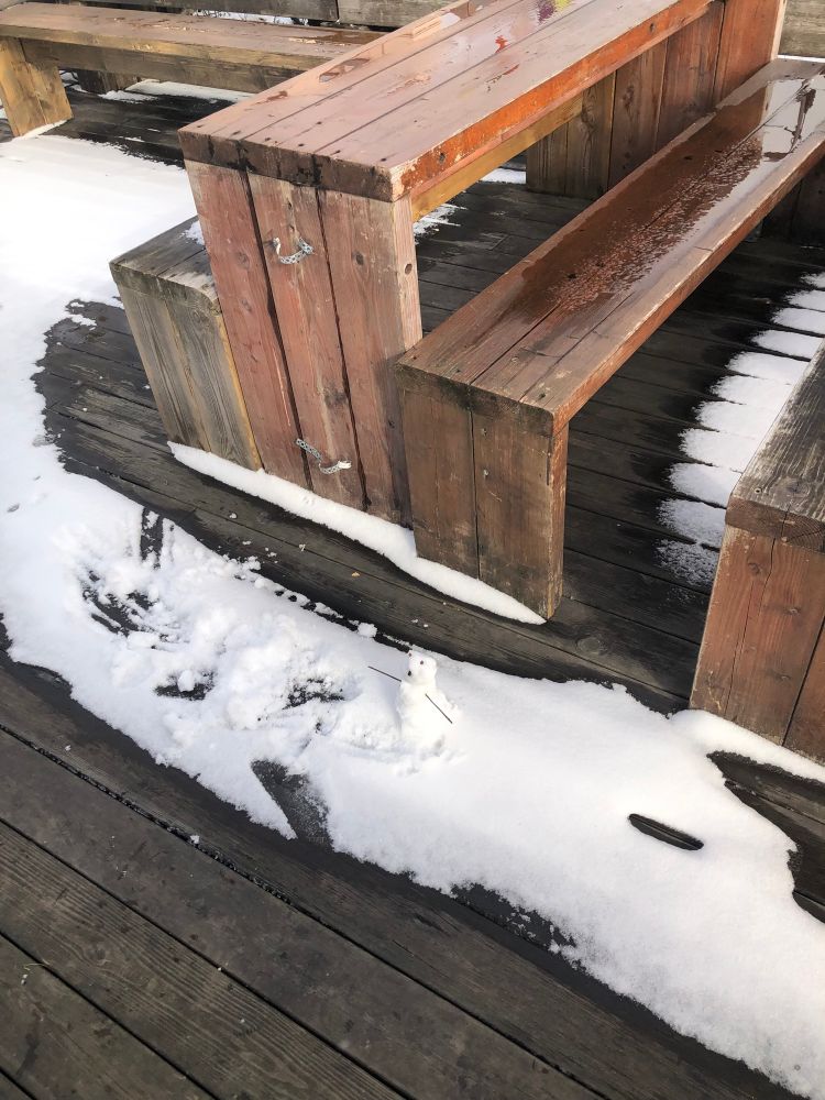 A very tiny snowperson sits in a small pile of snow next to a table. 