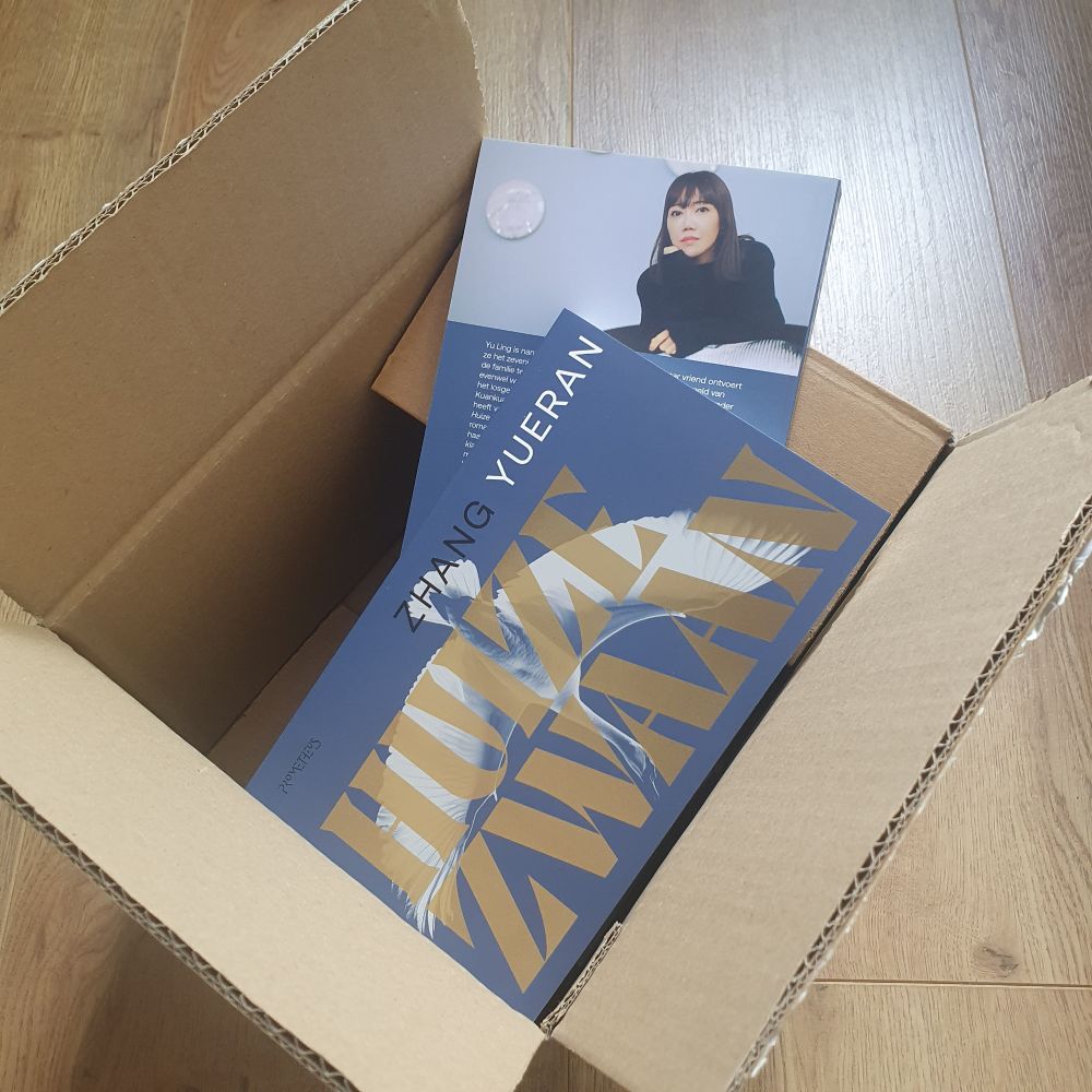 Foto van een open doos met daarin exemplaren van het boek Huize Zwaan van Zhang Yueran. Blauw omslag, gele letters, met achter de letters een vliegende zwaan.