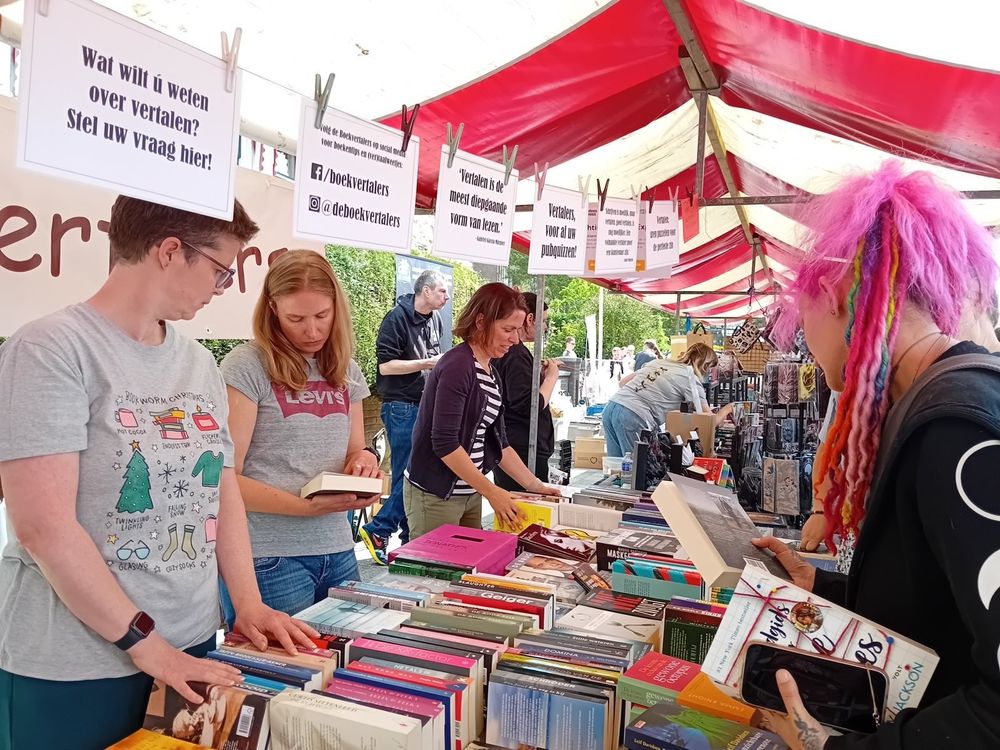 Foto van de Boekvertalers-kraam. Achter de kraam staan drie enthousiaste boekvertalers, voor de kraam is een vrouw met lang roze haar te zien die een van de boeken bekijkt. Door de kraam hangt een slinger met teksten, zoals 'Wat wilt u weten over vertalen? Stel uw vraag hier!' en 'Wat AI kan, kunnen wij beter'.