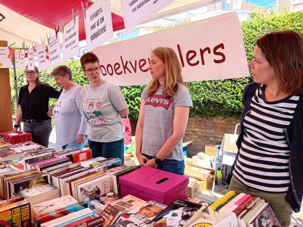 Foto van de Boekvertalers-kraam. Achter de kraam staan enthousiaste boekvertalers, met voor hen allemaal vertaalde boeken en achter hen een banner met 'Boekvertalers'. Door de kraam hangt een slinger met teksten, zoals 'Vertalers, voor al uw pubquizzen' en 'Wat AI kan, kunnen wij beter'.
