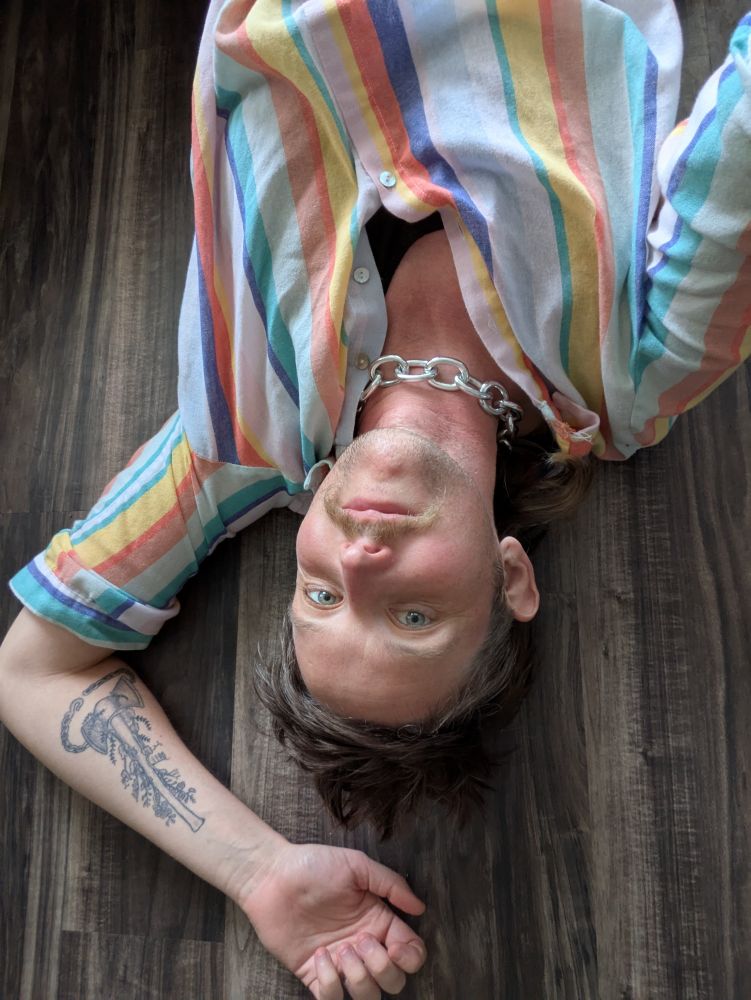 Middle aged white androqueer lies on the floor. Their shirt has coloured stripes. Their necklace looks like heavy chain. Their forearm tattoo of sweet peas climbing a labrys is visible. Their ice-green eyes look at the camera. 