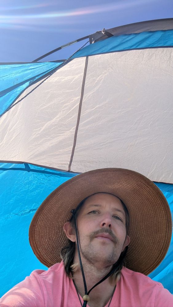 Middle aged scruffy faced white non-binary trans person sits under a sun shade, wearing a big sun hat and pink v-neck shirt. Rainbows dance in the wispy clouds above 