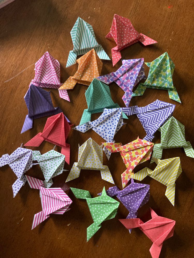 21 origami frogs are arranged on a wooden table.  Each frog is a different colour and pattern: red, yellow, green, pink, purple, striped, spotted, dotted, and zigzagged.