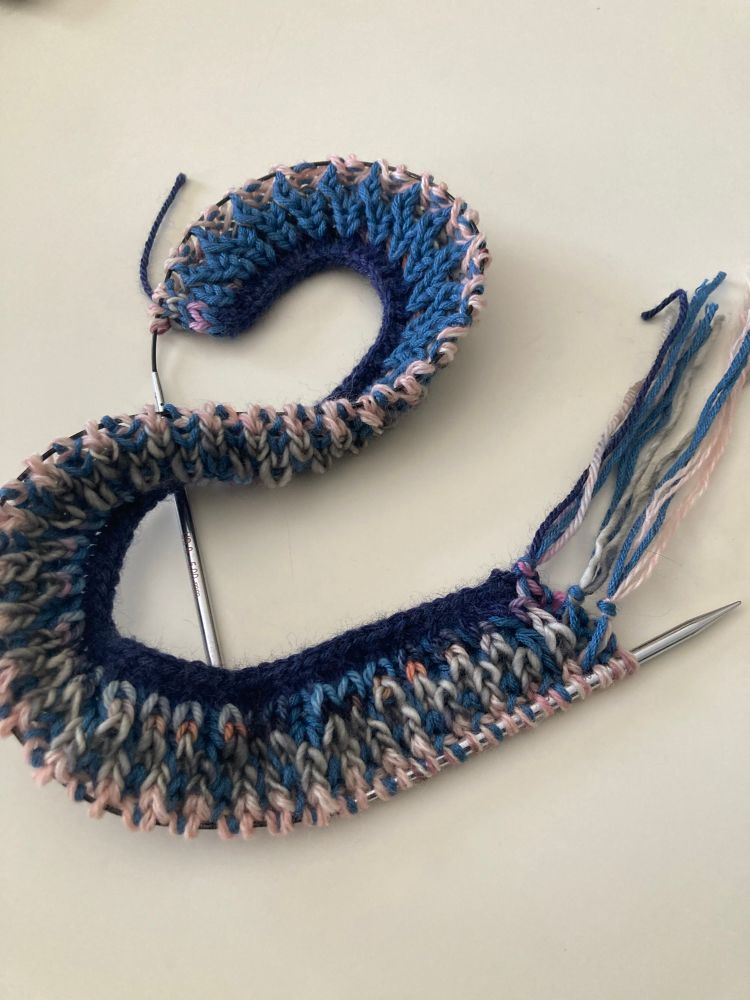 A multicoloured (mainly blues and soft pinks), brioche knitting project on a white background.  The knitting cable is twisted so you can see both sides of the few rows of knitting - which allows different colours to be dominant on each side.  One side is more blue and one side is more pink.  