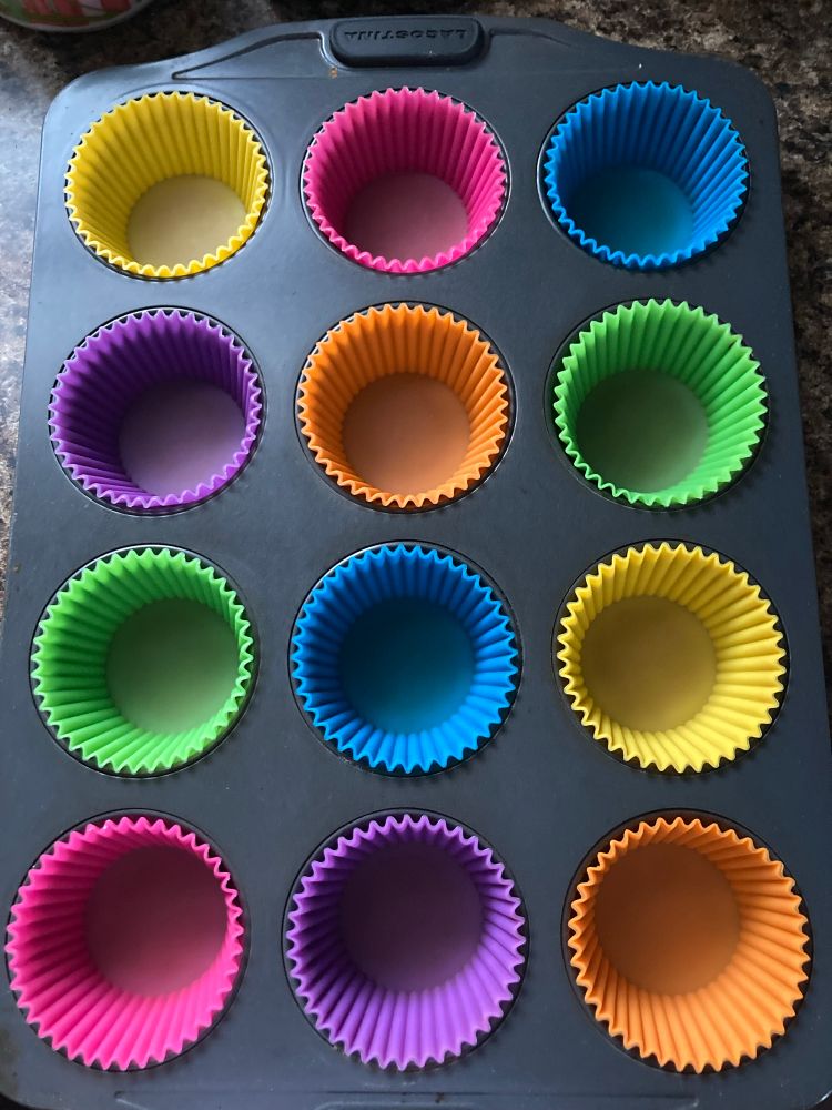 Neon coloured, silicone muffin tray liners are photographed from above in a dark grey metal muffin tin.  Pink, orange, yellow, green, blue, and purple.  The same colour never appears twice in a row or column or even diagonally touching. 