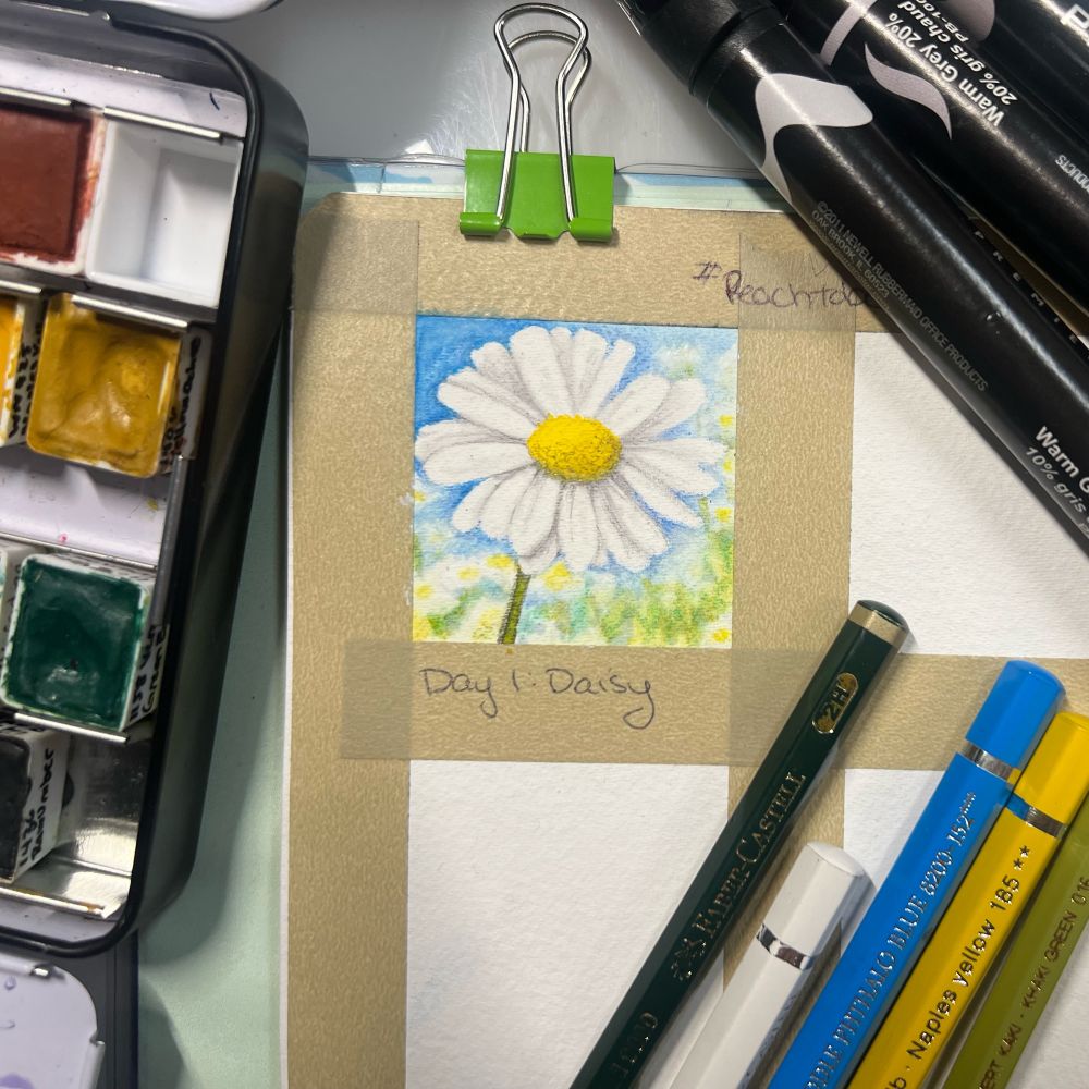 Up close photo of an open sketchbook with a small illustration of a daisy surrounded by pencils, markers and watercolor paint pans.