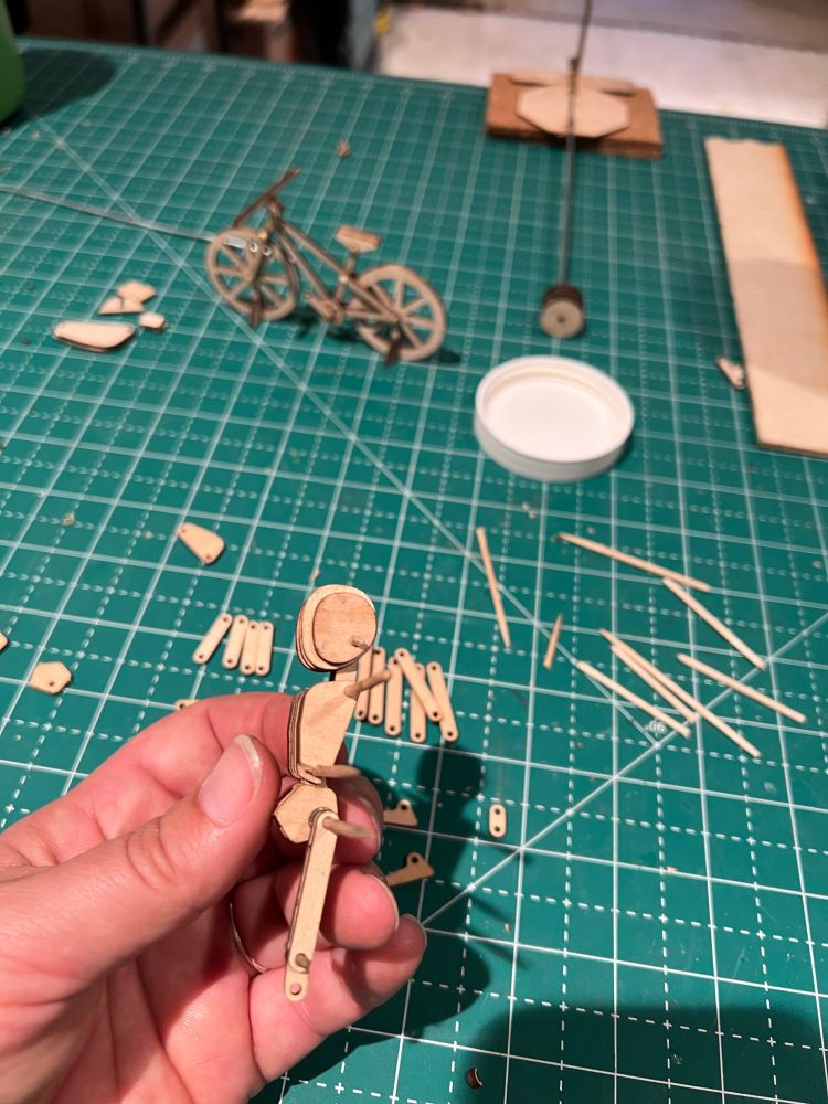 A photo of a hand assembling some wood cutouts to form a small human shaped figure. In the background is an already assembled bike made of similar wood pieces.