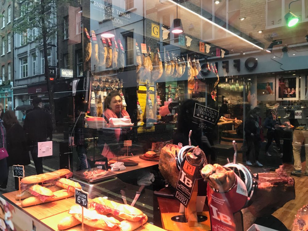 color photo shot through a shop window showing rows of hams hanging, sandwiches, a woman's face, and pedestrians reflected in the window, a windowed collage