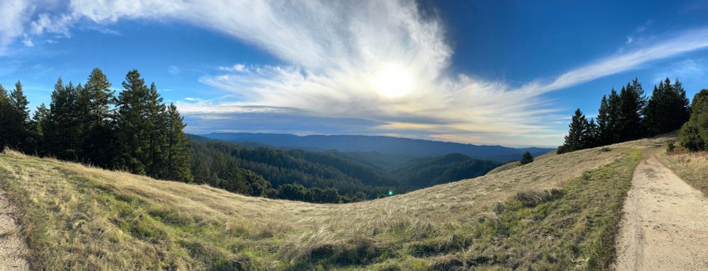 A panoramic view of a hilltop with bright blue sky, some clouds, and the ocean in the distance. 