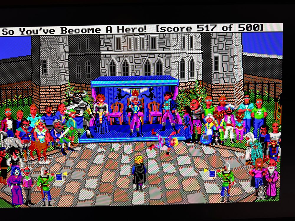 A photo of a computer monitor showing the ending of the video game Hero’s Quest, by Sierra Games. At the top it says “So You’ve Become A Hero! [score 517 of 500]”
The scene shows the hero standing in the courtyard of the castle surrounded by all the characters from the game, as well as some of the developers as Easter Eggs. 