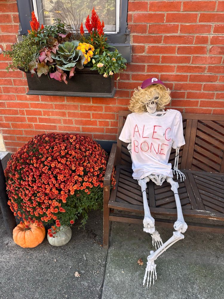 A prop skeleton sitting on a bench in front of a brick home. The skeleton is wearing a white T-shirt that reads “Alec Bone”. The skeleton is wearing a wig of curly blond hair, and a red Phillies cap.  There are lots of flowers and a pumpkin near the bench. 
