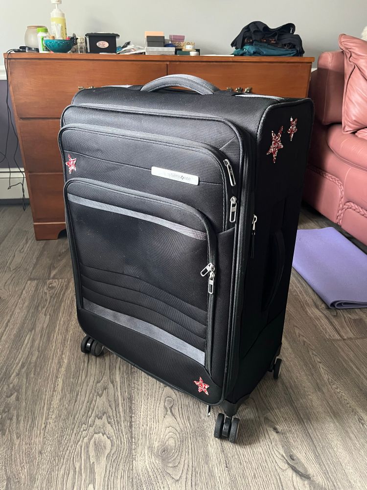 Picture of a black fabric Samsonite suitcase upright in three-quarters profile in front of a brown wood dresser.  There are 4 red and silver sparkly stars on the suitcase - 2 on the front in opposite corners (upper left and lower right), and 2 near the top of the side, one larger than the other.