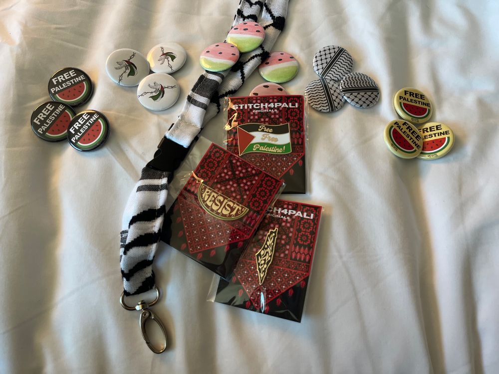 A Kuffiyeh fabric lanyard surrounded by 5 designs of button-style pins that show Palestinian Support, plus 3 designs of enamel pins still in packaging.
