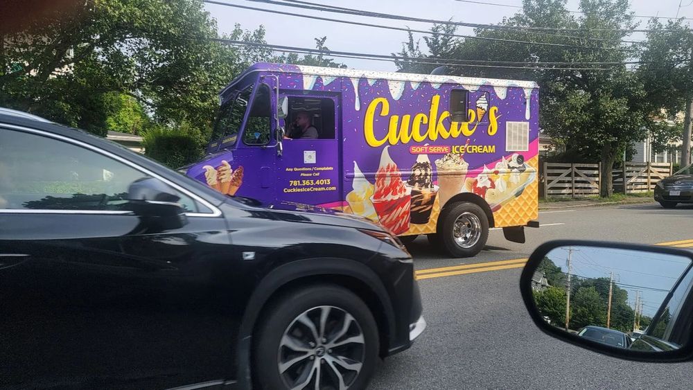 A picture out the side of a stopped car.  On the right is the rearview mirror.  On the left is a black sedan. In the center is a squarish truck painted purple with images of various ice creams on it, with the name “Cuckie’s” in yellow paint.