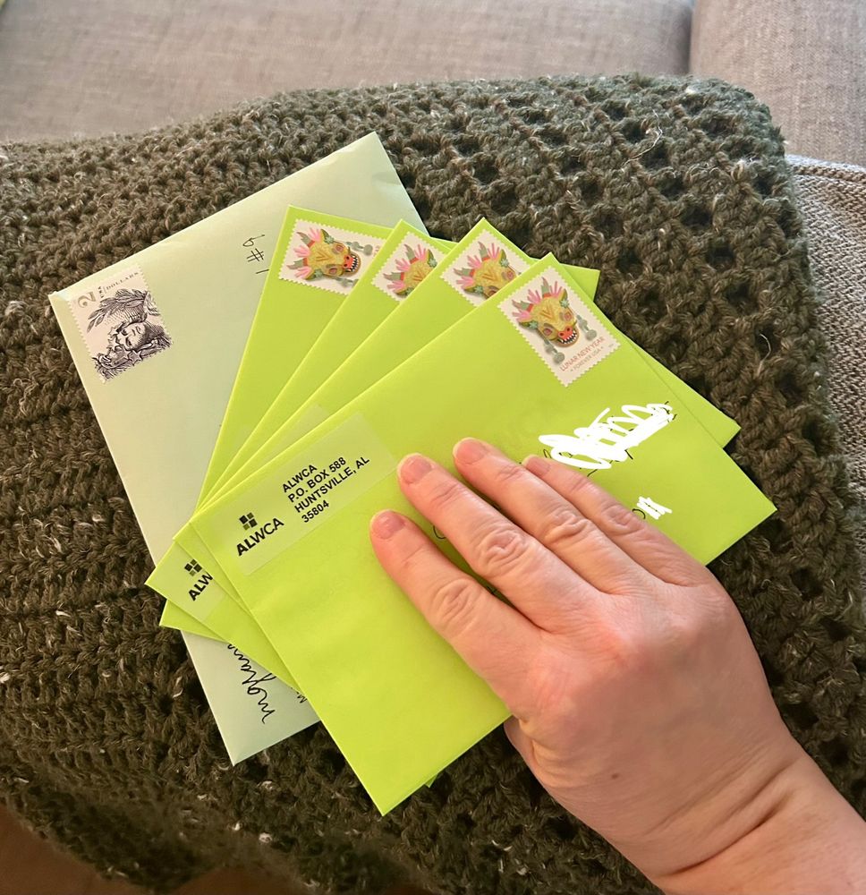 A photo of my right hand holding five envelopes. One envelope is bigger and light green. The other four are lime green. I am holding them in front of my green crocheted reading shawl folded over the back of my living room chair. 