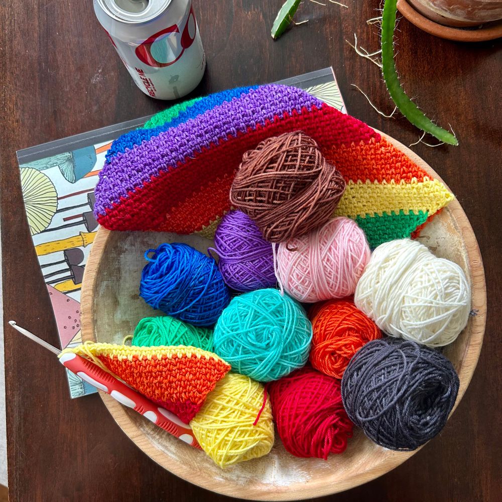 An old wooden bowl holding small skeins of yarn in the 11 Progress Pride flag colors. One finished square piece of crochet in the original rainbow colors is hanging out of the top of the bowl. The second square is in progress on a crochet hook in the bottom left of the bowl. The bowl sits on my rainbow mushroom notebook on my dark wood dining table. There’s an open can of Diet Coke in the upper left of the pic. There’s two cactus arms photobombing the scene from the top right.