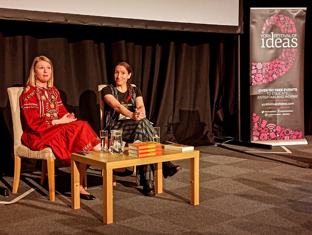 A wider shot of Dr Charlotte Proudman and Charlotte O'Brien sitting on chairs talking on a stage during a talk at York Festival Of Ideas. They are both looking into the audience. There is a small table in front of them, with copies of Dr Proudman's book, and a jug and glasses of water. There is also a banner on the far right side, promoting the York Festival Of Ideas.