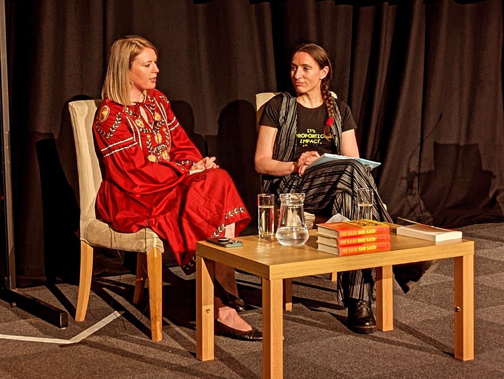 Dr Charlotte Proudman and Charlotte O'Brien sitting on chairs talking on a stage during a talk at York Festival Of Ideas. There is a small table in front of them, with copies of Dr Proudman's book, and a jug and glasses of water.