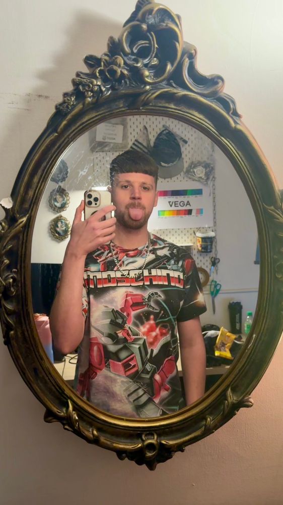 a picture of Danny (mushy) framed in the reflection of an oval shaped mirror. behind him on the wall is a board full of colour swatches for the Vega Workshop harnesses.