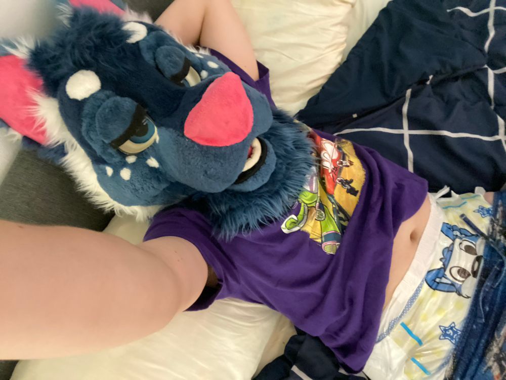 A person wearing a blue, white, and pink fursuit head and a purple t-shirt is lying down on a bed. The fursuit head features large, bright blue eyes, a pink snout, and prominent ears with pink inner ear details. The person's arms are visible, with one arm resting above their head. A Peek-abu diaper with a blue cartoon husky pokes out from above their waistband