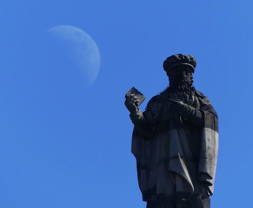 Foreground: a C19th statue of John Knox, wearing a puffy hat and holding up what is presumably a Bible but looks very much like his lunch. Background: the Moon, clearly visible in a blue sky.
