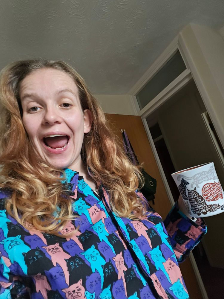 Rosie wearing a bright, long sleeved shirt with hundreds of cats on it while drinking a hot chocolate from a mug with cat quotes on