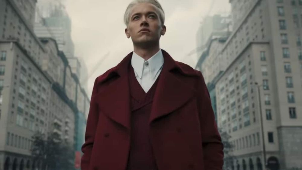 Coriolanus Snow at age 18, wearing a white shirt and red coat, standing with a view of two sides of a city street behind him, looking forward and up with a cold gaze.