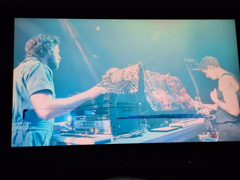 Three men around an electronic music console at a concert stage. There us a bright light with blue hues shining on the console.