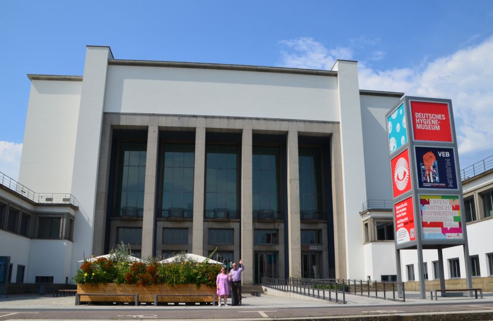 Photo of the main entrance to the "Deutsches Hygiene-Museum" in Dresden. In the foreground, very small, two people waving. Members of the organising teams of the IOER Conference 2024: Jessica Hemingway on the left, Wolfgang Wende on the right.