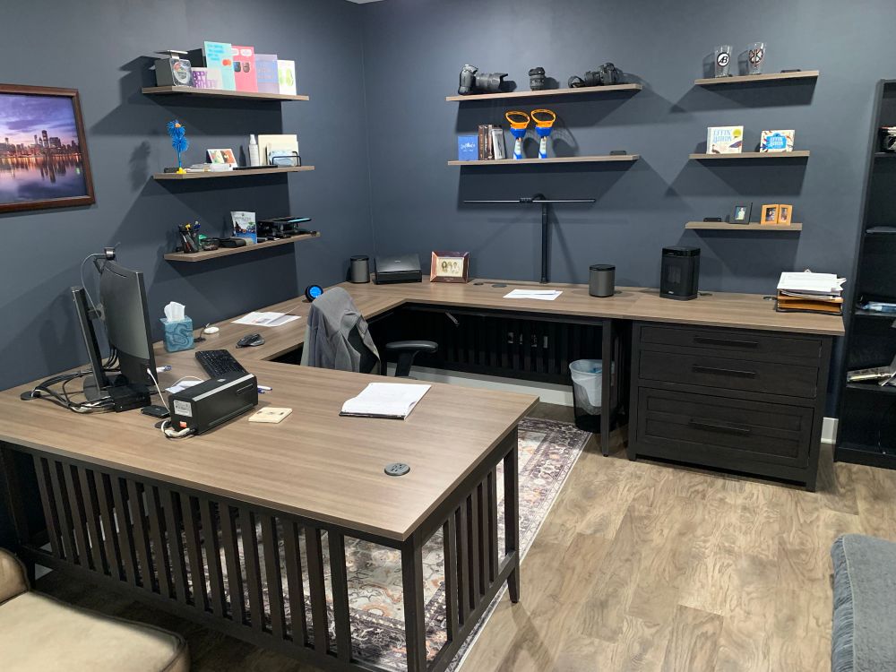 A home office with U shaped desk, drawer unit, 8 floating shelves, and dark blue/grey walls. 