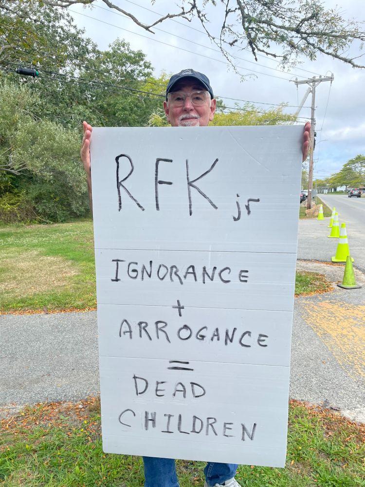 Some old dude (me) wearing a baseball cap holds a large hand-written sign: "RFKjr  IGNORANCE + ARROGANCE == DEAD CHILDREN