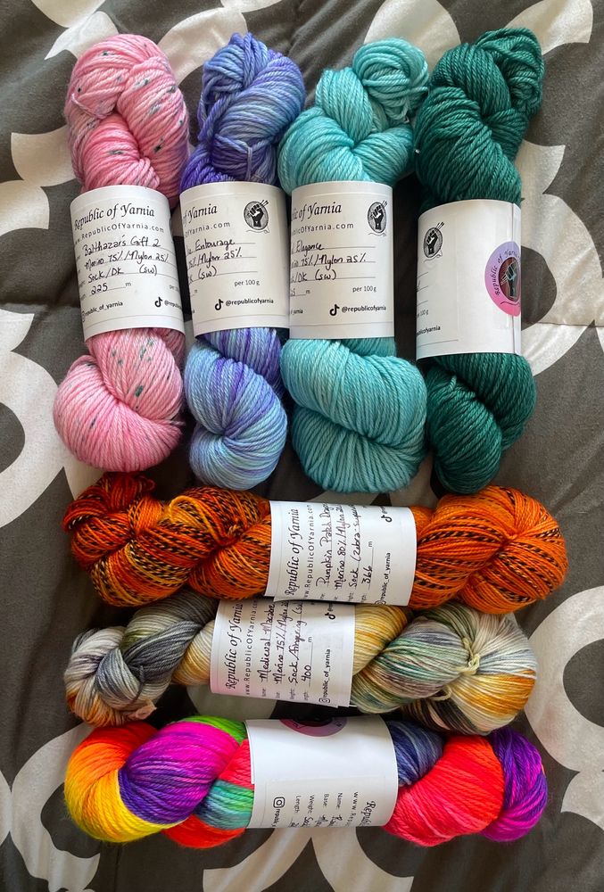 7 hanks of yarn: speckled pink, pale blue/purple, a light teal, a dark green teal, a bright orange with black marled stripes, a white-grey-orange, and finally a ridiculously bright neon rainbow 