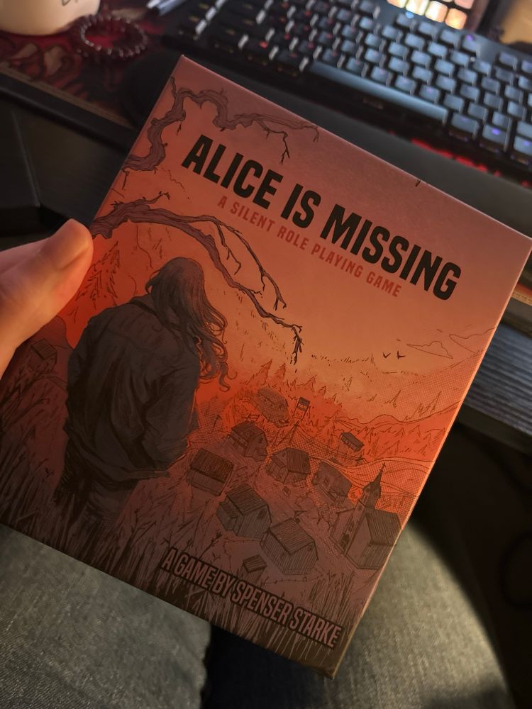 Physical copy of Alice Is Missing: A Silent Roleplaying Game by Spenser Starke. Box shows an individual with long hair looking down on a town painted in pinks and reds.