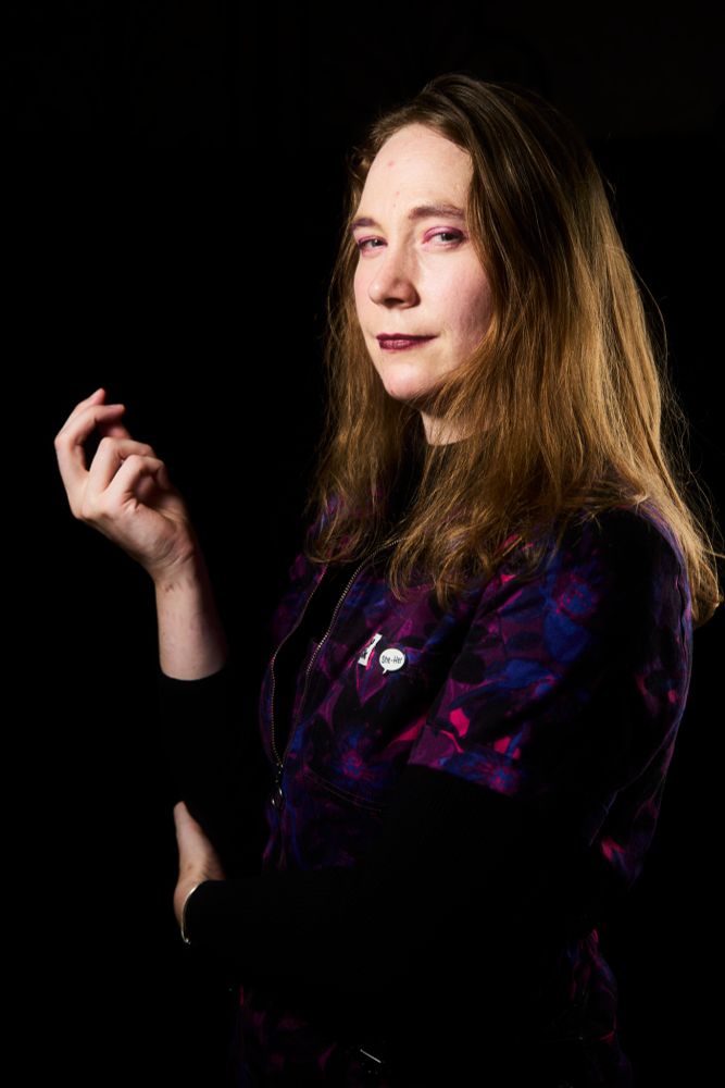 Fiona, a white woman with shoulder length blonde hair wearing a purple patterned jumpsuit from Lucy & Yak in a side on profile, holding her right hand up in a thoughtful gesture. Headshot by K11 Studios.