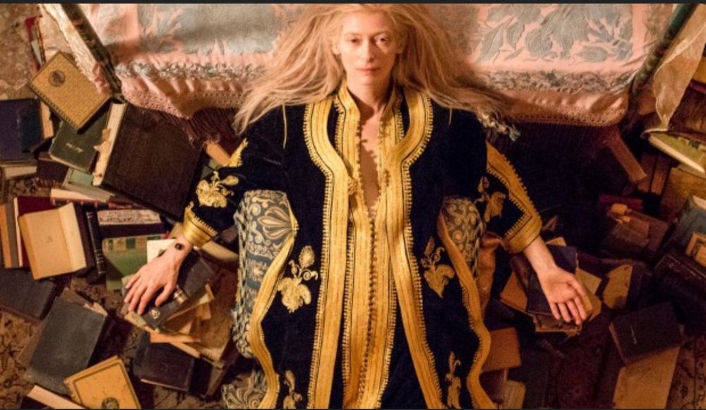 An image taken from above of a pale, blonde-haired, dark-eyed woman in a luxurious long black and gold outfit lying on on her back on a bed. She is surrounded by old hard copy books. One palm rests on a book, the other is open and upwards.