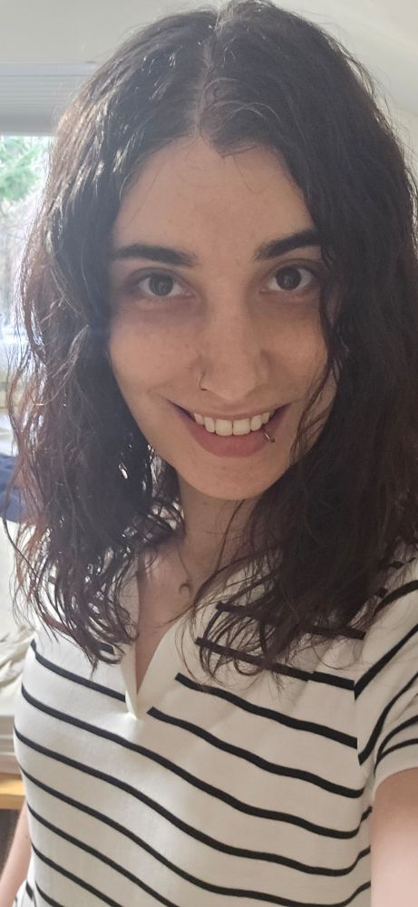 Selfie of a woman with shoulder length wavy brown hair wearing a black and white striped dress and a crescent moon necklace 