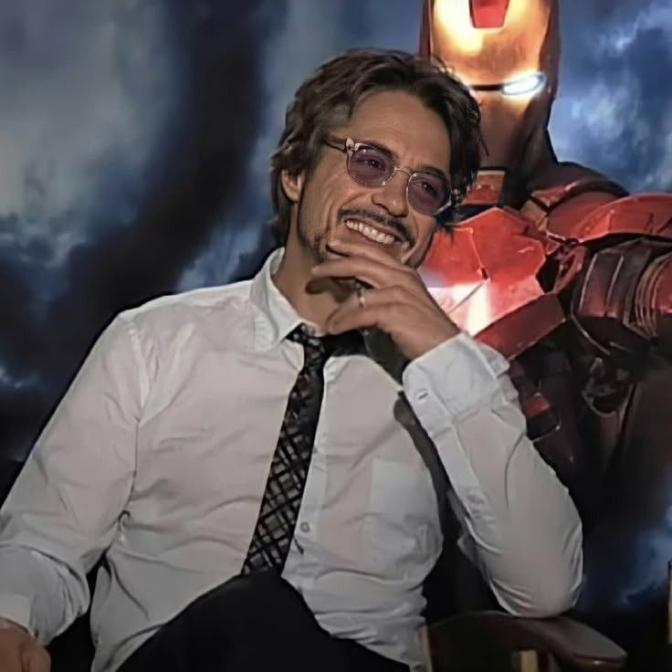 robert downey jr on one of the press tours for iron man. he's in most likely an interview, with the film cover / iron man imagery in the background. he's wearing pink tinted sunglasses, a patterned tie, a white button down, and black pants. he's smiling and looking at something ofd camera. his finger is against his jaw and his elbow is resting on the chair, posing. 