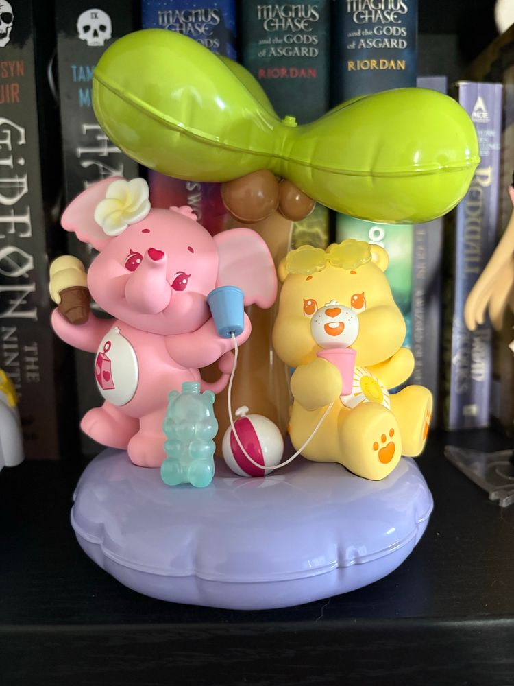 A figure of Lotsa Heart Elephant and Funshine Bear. They are on a purple inner tube with an inflatable coconut tree sprouting from the center. Lotsa stands with an ice cream cone and tin can phone in each hand. Funshine sits on the inner tube, talking to the tin can phone in their hand